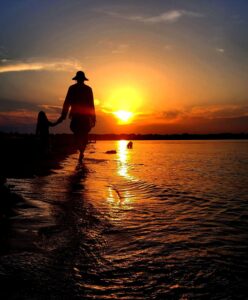 Erwachsene Person und Kind gehen Hand in Hand am Wasser in den Sonnenuntergang, als Silhouetten